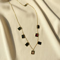 A gold chain necklace with multiple rectangular pendant stones in black, red, and smoky brown, arranged on draped cream-colored fabric.