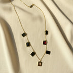 A gold chain necklace with multiple rectangular pendant stones in black, red, and smoky brown, arranged on draped cream-colored fabric.