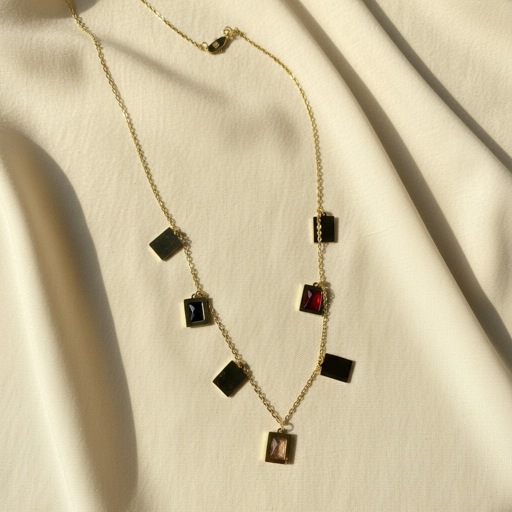 A gold chain necklace with multiple rectangular pendant stones in black, red, and smoky brown, arranged on draped cream-colored fabric.
