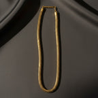 A gold-toned snake chain necklace with a polished bar clasp, displayed on draped dark gray fabric.