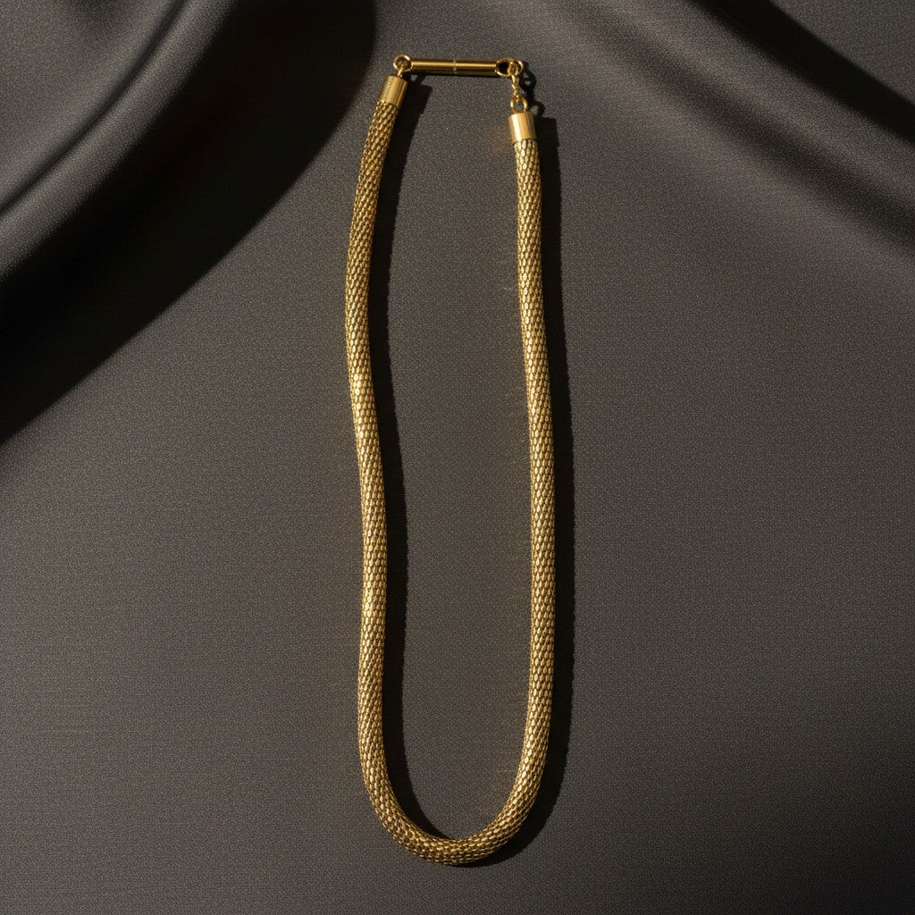 A gold-toned snake chain necklace with a polished bar clasp, displayed on draped dark gray fabric.
