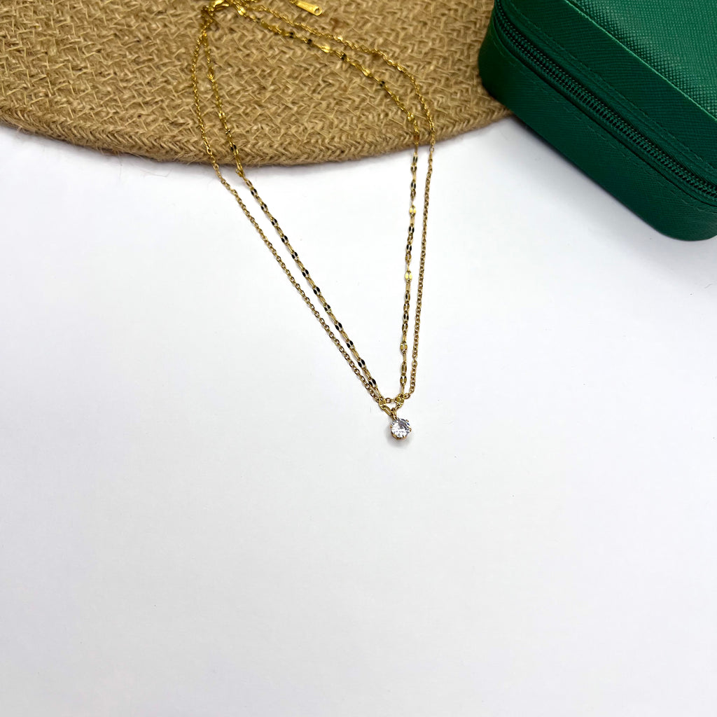 Layered 18K gold plated chain with a solitaire CZ pendant placed on a white background beside a wooden prop

