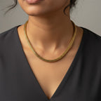 A close-up of a model wearing a delicate gold chain necklace with a textured, woven mesh-like appearance, styled with a black V-neck top against a neutral gray background.