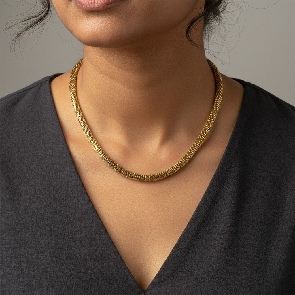 A close-up of a model wearing a delicate gold chain necklace with a textured, woven mesh-like appearance, styled with a black V-neck top against a neutral gray background.